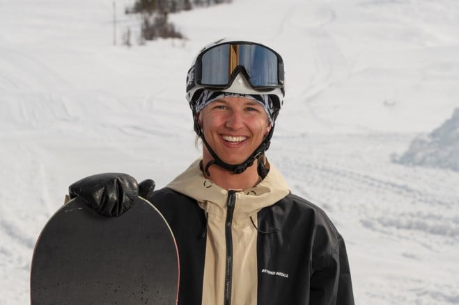 Smilende gutt med snøbrett i hånda og alpinbakke i bakgrunnen